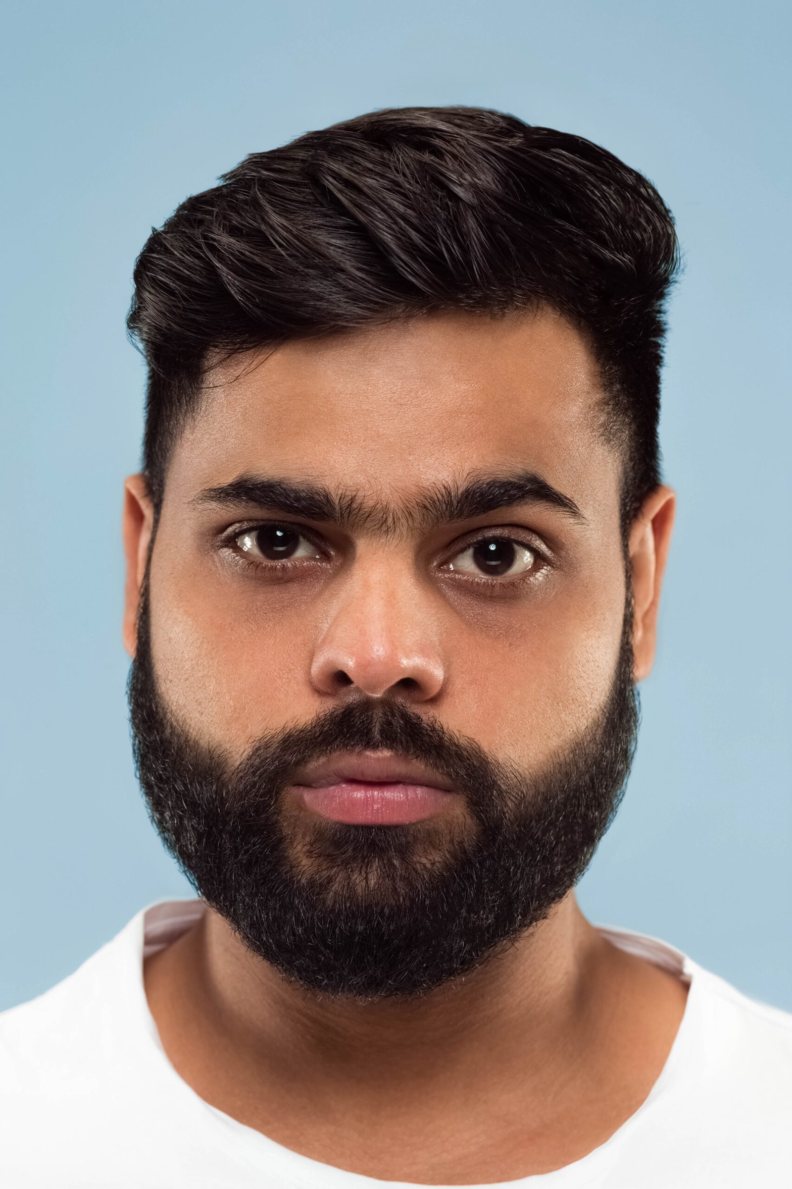 Close up portrait of young hindoo man with beard in white shirt isolated on blue background. Human emotions, facial expression, ad concept. Negative space. Standing and looking calm.